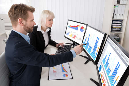 Two Young Businesspeople Analyzing Graph On Multiple Computer At Officeの写真素材