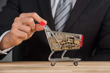 Close-up Of Businessman Carrying Coin In Shopping Trolley At Wooden Deskの写真素材