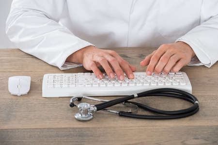 Close-up Of Male Doctor Typing On Computer Keyboard With Mouse At Wooden Desk In Clinicの写真素材
