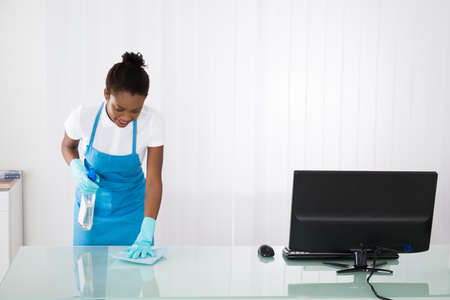 Happy Young African Female Janitor Cleaning Desk With Rag In Office