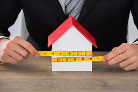 Close-up Of Businessman Hand Measuring House Model At Wooden Deskの写真素材