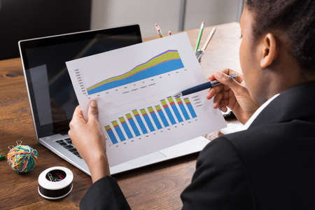 Close-up Of African Businesswoman Analyzing Graph With Laptop On Wooden Deskの写真素材