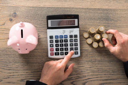 Close-up Of A Businessman Counting Coins Using Calculator At Desk In Office