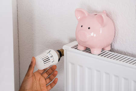 Close-up Of Woman's Hand Adjusting Thermostat With Piggy Bank On Radiatorの写真素材