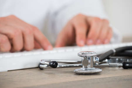 Close-up Of Male Doctor Using Computer Keyboard With Stethoscope On Wooden Desk In Clinicの写真素材