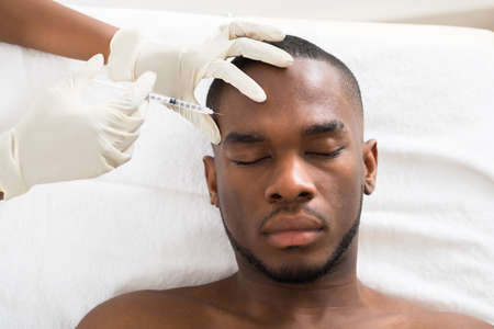 Close-up Of Person Hand Injecting Syringe On Young African Man Face