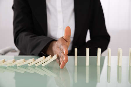 Close-up Of Businesswoman Hand Stopping Dominoes From Falling On Office Deskの写真素材