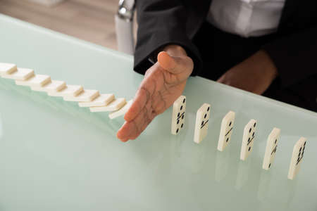 Close-up Of Businesswoman Hand Stopping Dominoes From Falling On Office Deskの写真素材