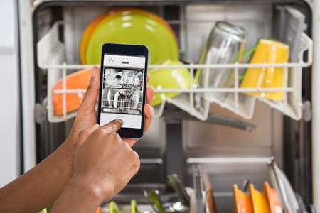 Close-up Of Person Hands Operating Dishwasher With Mobile Phoneの写真素材