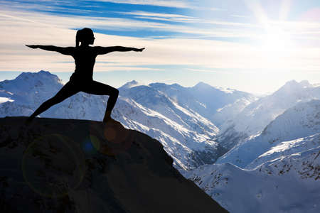 Silhouette Of A Woman Practicing Yoga On A Cliff Against Snowy Mountainsの写真素材
