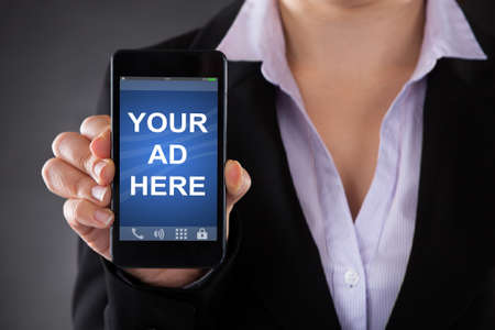 Close-up Of Person Showing Your Ad Here Text On Mobile Phone Against Grey Background