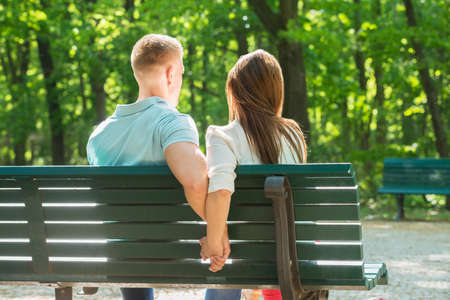 Rear View Of Couple Sitting On Bench Together And Holding Hands At Parkの写真素材