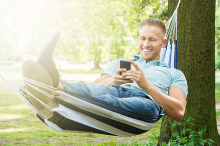 Young Happy Man Using Mobile Phone While Relaxing In Hammock At Gardenの写真素材