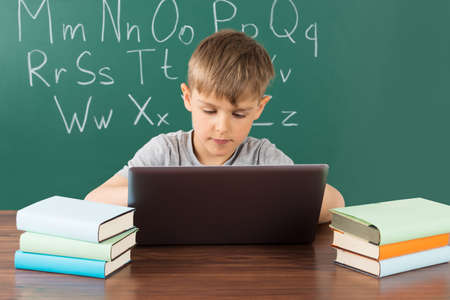 Boy Using Laptop In Front Of Green Chalkboard At School