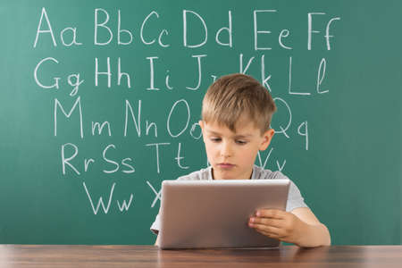 Boy Using Digital Tablet In Front Of Green Chalkboard At School