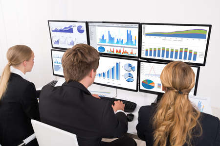 Young Male And Female Businesspeople Looking At Financial Graphs On Multiple Computers In Office
