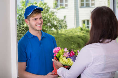 Smiling Young Woman Receiving Bouquet Of Flowers From Delivery Man At Homeの写真素材