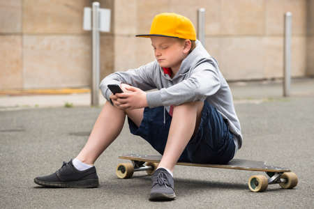 Boy Wearing Cap Sitting On His Skateboard Using Smartphoneの写真素材