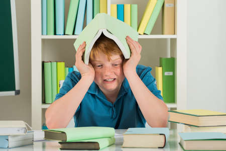 Angry Boy Sitting In Library Holding Book On His Head
