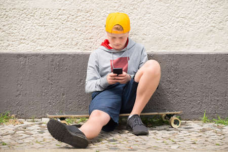 Boy Sitting On Skateboard Using His Mobile Phoneの写真素材