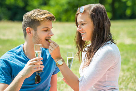 Woman Feeding Grape To Her Boyfriend With Glass Of Champagne In Handの写真素材