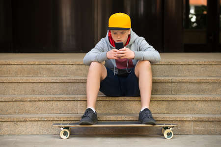 Boy Wearing Cap Sitting On Staircase Looking At Cellphoneの写真素材