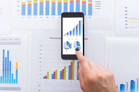 Close-up Of Businesswoman Analyzing Financial Graphs Using Smartphone On Deskの写真素材