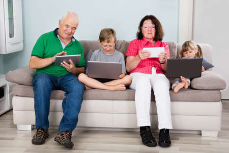 Family With Kids Sitting On Couch With Laptop Computers And Tablets At Homeの写真素材