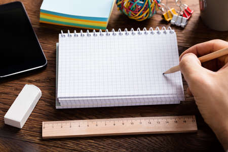 Close-up Of Person Hand Holding Pencil On Blank Notepad Near Scale And Eraser At Wooden Deskの写真素材