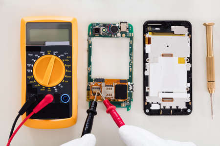 High Angle View Of Person Checking Damaged Mobile Phone With Digital Multimeter