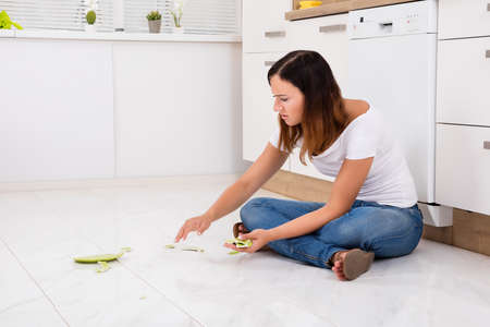 Young Unhappy Woman Looking At The Broken Plate Fallen On Floor In The Kitchenの写真素材