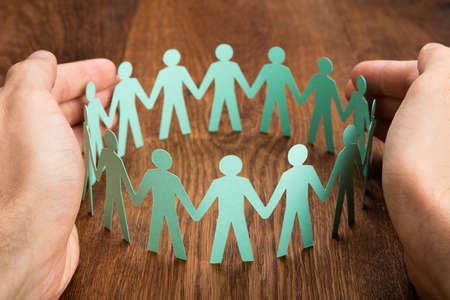 Close-up Of Person Hand Protecting Cut-out Figures On Wooden Desk