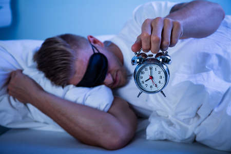 Man Sleeping On Bed By Holding Alarm Clock In His Hand
