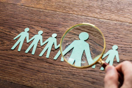 High Angle View Of Person Hand Using Magnifying Glass On Cut-out Figures On Wooden Desk