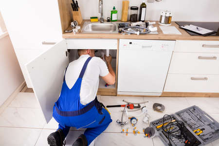 Male Plumber In Overall Repairing Sink Pipe In Kitchen