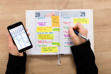High Angle View Of A Businesswoman Writing Schedule In Diary Using Mobilephone On Wooden Desk