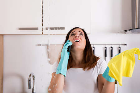 Young Shocked Woman Calling To Plumber While Water Leaking From Ceiling In Kitchenの写真素材