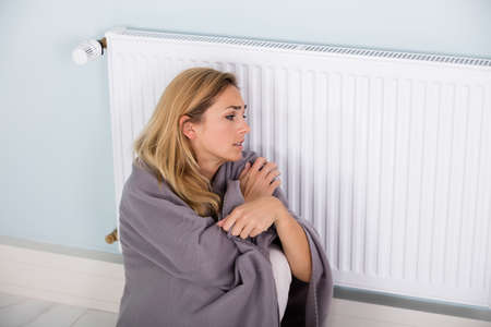 Young Cold Woman Wrapped In Blanket Sitting Near Central Heating System At Homeの写真素材