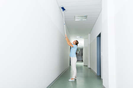Young Woman Cleaning The Ceiling With Mop On The Corridor Of The Building