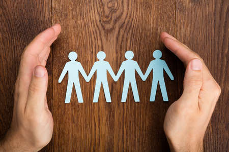 Close-up Of Person Hand Protecting Human Figure Cutout On Wooden Deskの写真素材