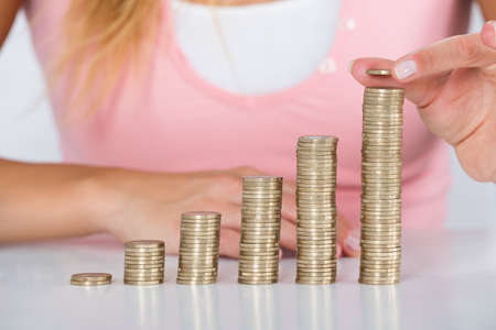 Close-up Of Woman Hand Inserting Coin On The Stack Of Coins At White Deskの写真素材
