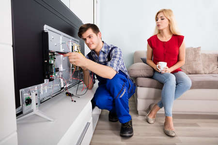 Young Woman Sitting On Couch Looking At Male Technician Repairing TV At Homeの写真素材