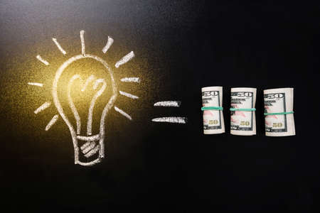 Equal Sign Between Light Bulb And Rolled Currency Note On Blackboardの写真素材