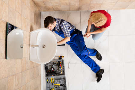 Woman Looking At Young Male Plumber Lying On Floor Fixing Sink In Bathroomの写真素材