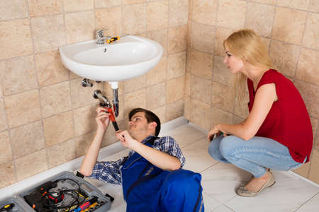 Woman Looking At Young Male Plumber Repairing Sink In Bathroomの写真素材