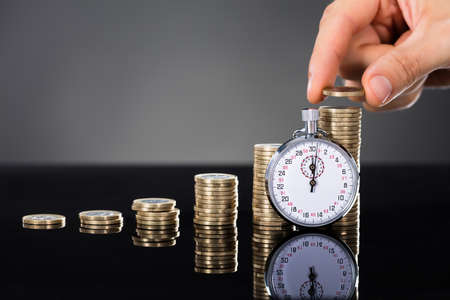 Close-up Of A Person Stacking Coins On Desk Near Stopwatch Over Gray Background