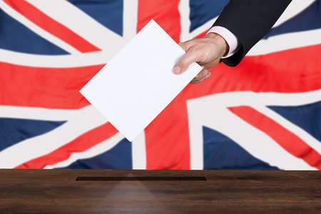 Businessperson Hand Putting Vote Into Ballot Box In Front Of United Kingdom Flag