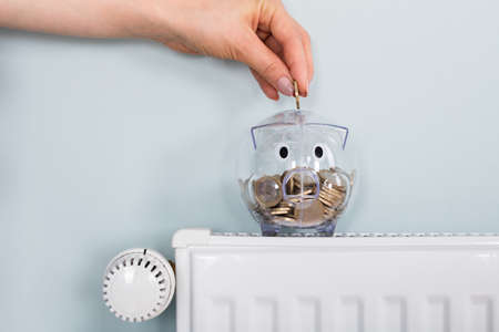 Close-up Of Person Hand Inserting Coin In Transparent Piggybank Kept On Radiatorの写真素材