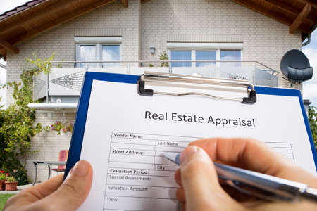 Close-up Of Person Hand Filling Real Estate Appraisal Document In Front Of Houseの写真素材