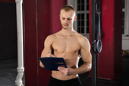 Portrait Of An Athlete Man Writing A Workout Plan On Clipboard In The Gymの写真素材